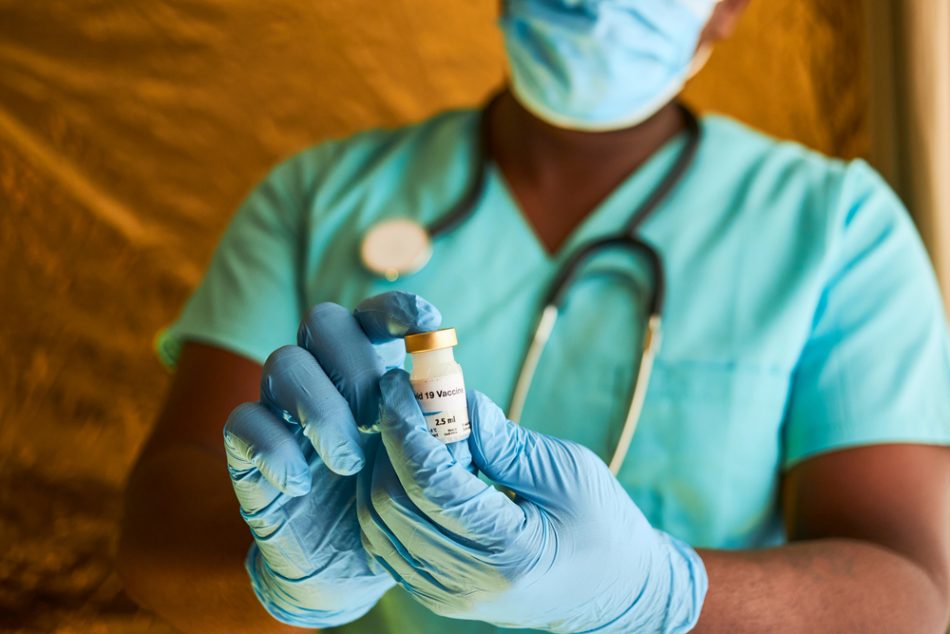 An African doctor holding a covid 19 vaccine vial in his hands while wearing blue surgical gloves and surgical scrubs.