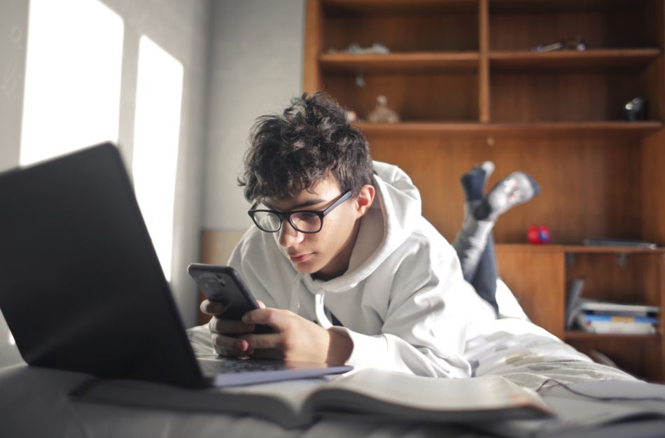 Young boy studies lying on the bed using computer and smartphone.