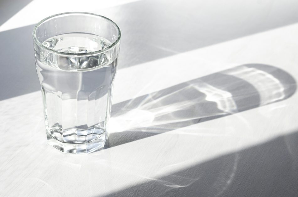 A glass with clean clear water and sharp shadows stands on a white wood table.