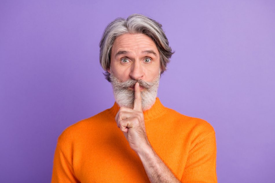 Photo of aged man in orange sweater i front of a purple background with finger over lips shh to stop interrupting.