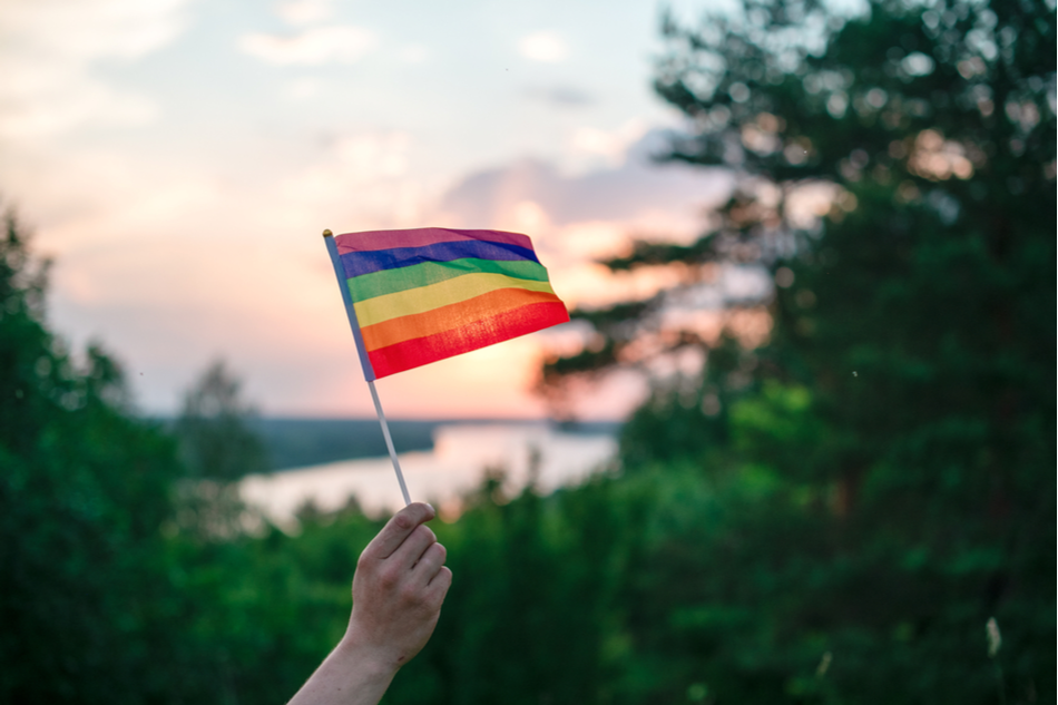 Hand holds LGBTQ rainbow flag against green and natural backdrop