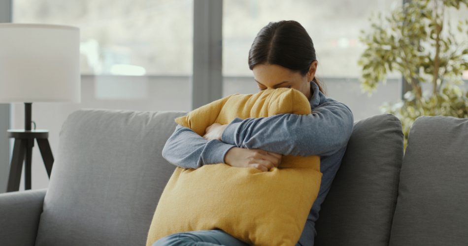Woman in a blue shirt hugging a yellow pillow on a grey couch.