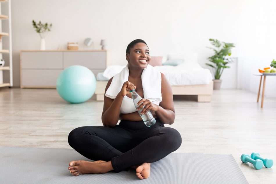Woman exercising on a sports mat at home drinking water with a towel round their neck.