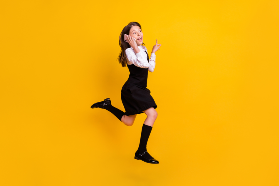 Little girl jumps happily while talking on the phone against a mustard yellow background