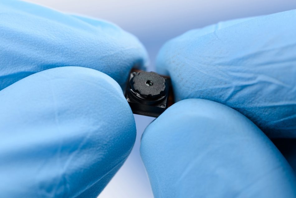 Tiny camera held between the fingers of a scientist wearing blue gloves.