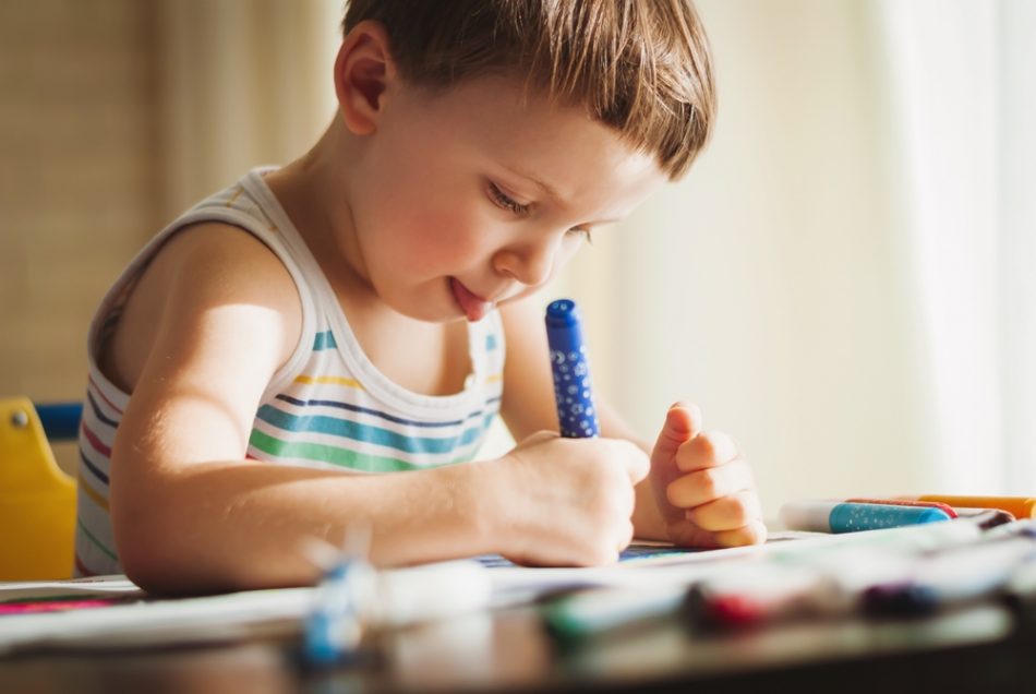 Small child sitting at a table and coloring sticking their tongue out in concentration.
