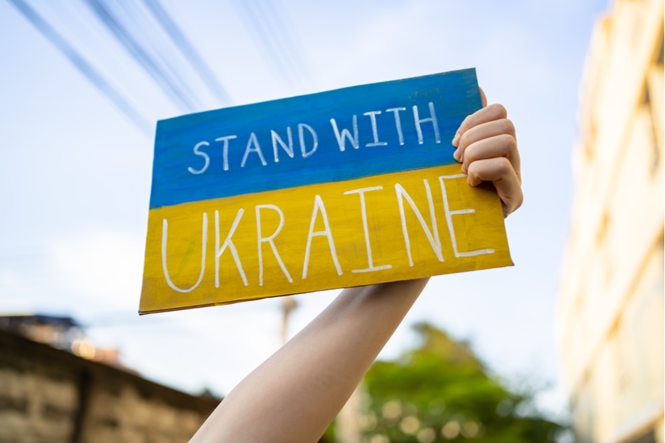 hand holds sign depicting Ukrainian flag with words: stand with Ukraine