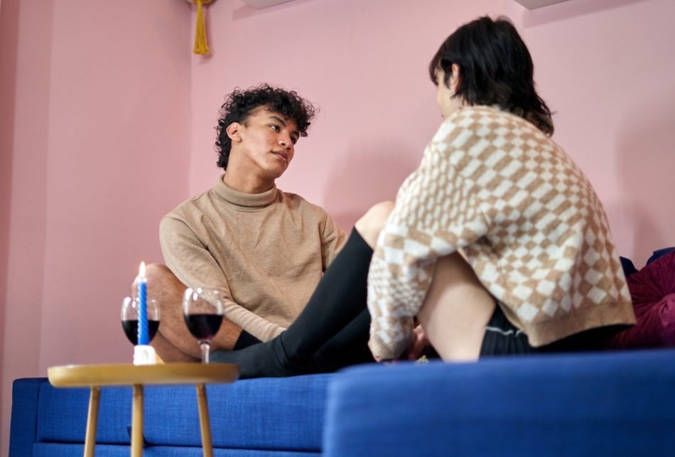 Male couple talking at home on blue couch with glasses of wine.