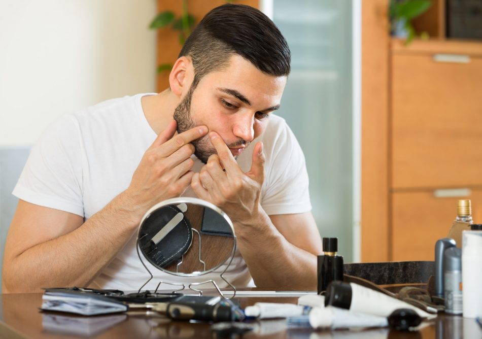 Young man pops pimple while looking into magnifying mirror