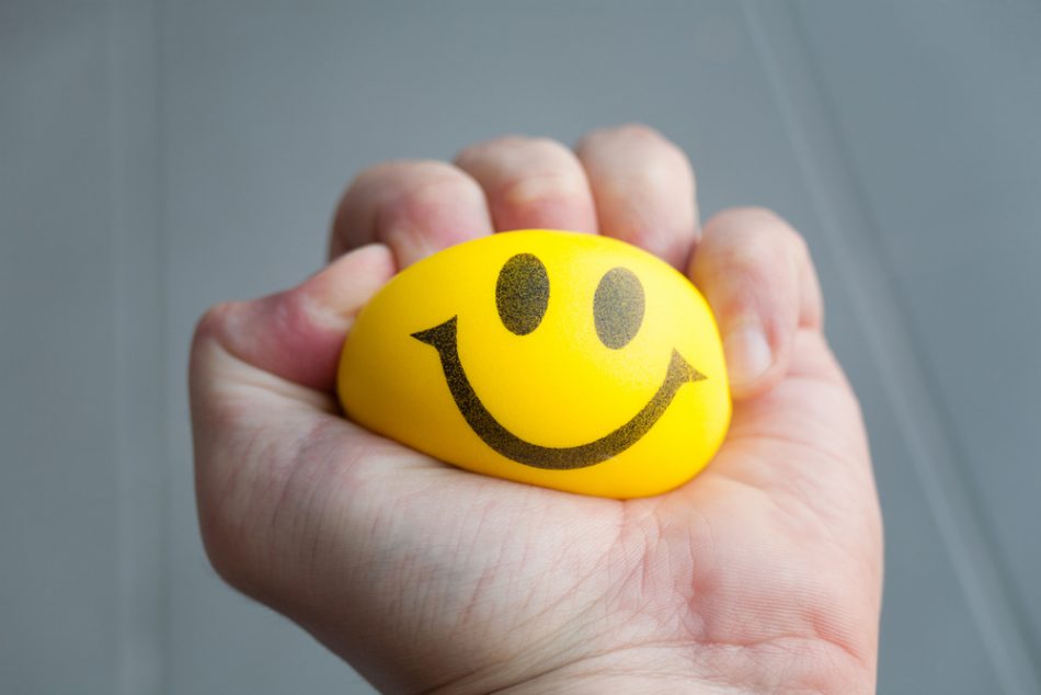 Person's hand squeezing a yellow stress ball with a smiley face on.