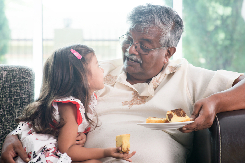 Indian grandfather sits with grandchild on the couch while eating cake