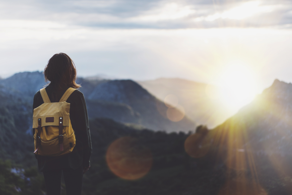 a female traveler's back as she views the sun through a mountainous landscape