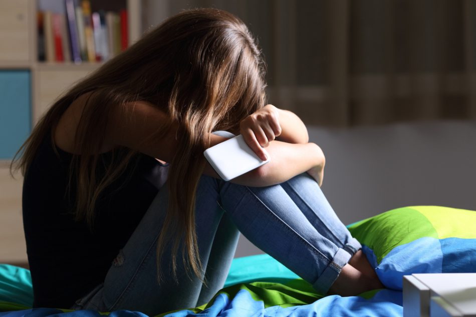 Single sad teen holding a mobile phone lamenting sitting on the bed in her bedroom with a dark light in the background representing teen anxiety.