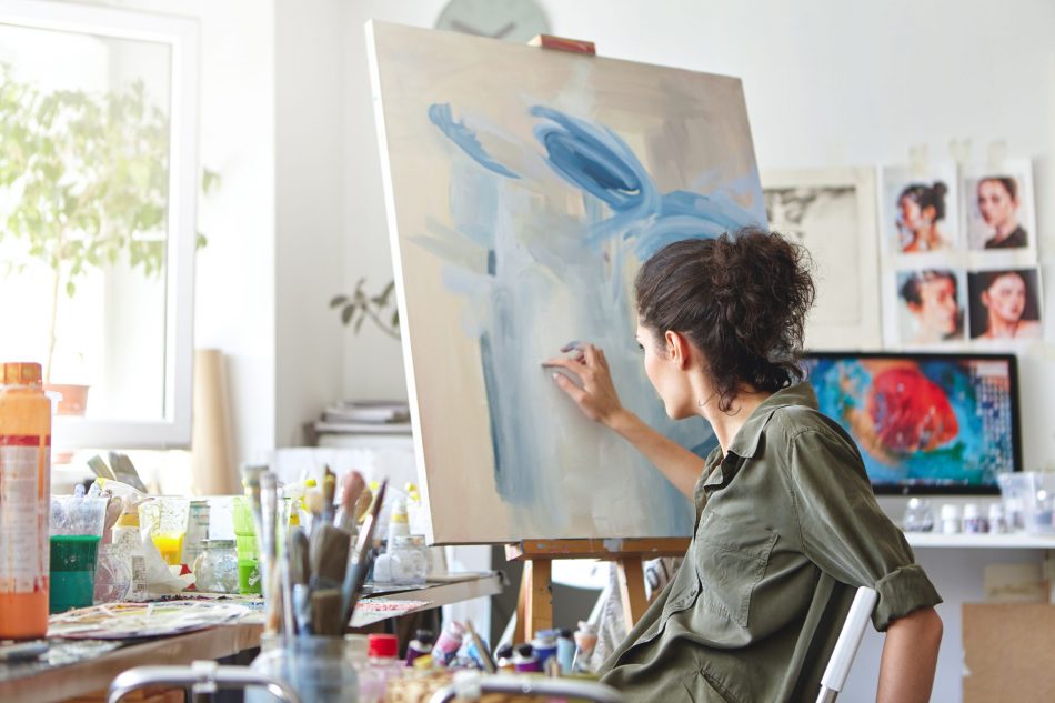 Rear view of busy female artist sitting on chair in front of easel, painting with fingers, using white and blue oil or acrylic paint.