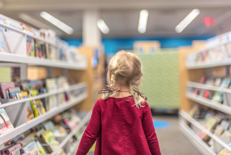 Little blonde girl walks through book shelves at library