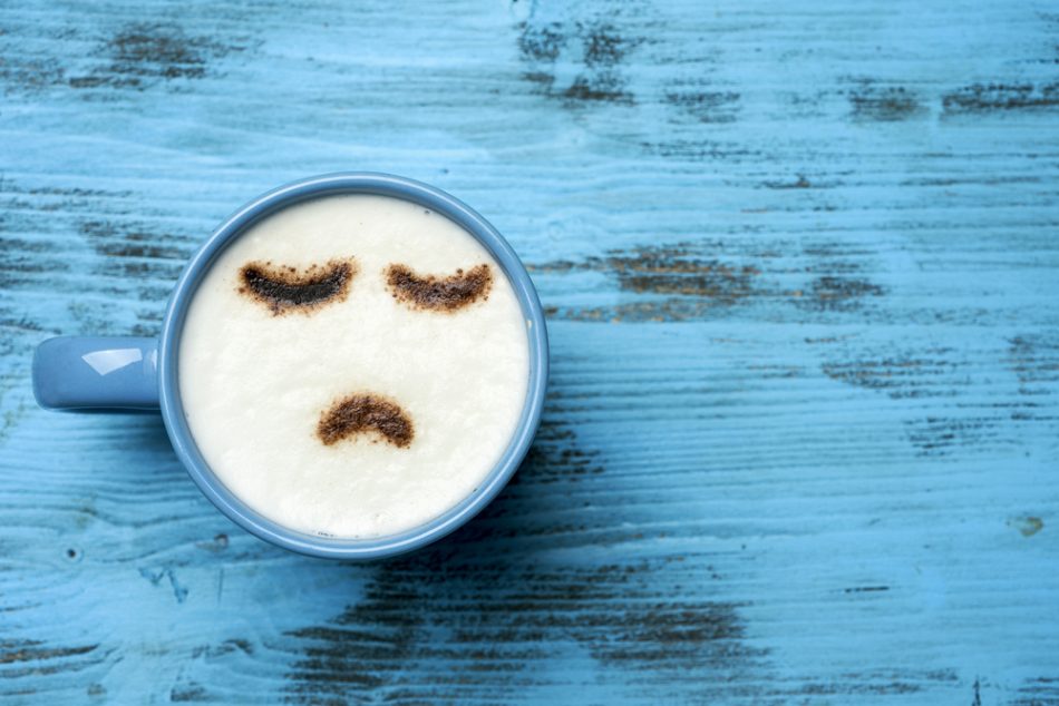 High-angle shot of a blue cup of cappuccino with a sad face drawn with cocoa powder on its milk foam, on a blue rustic table with a blank space on the right.