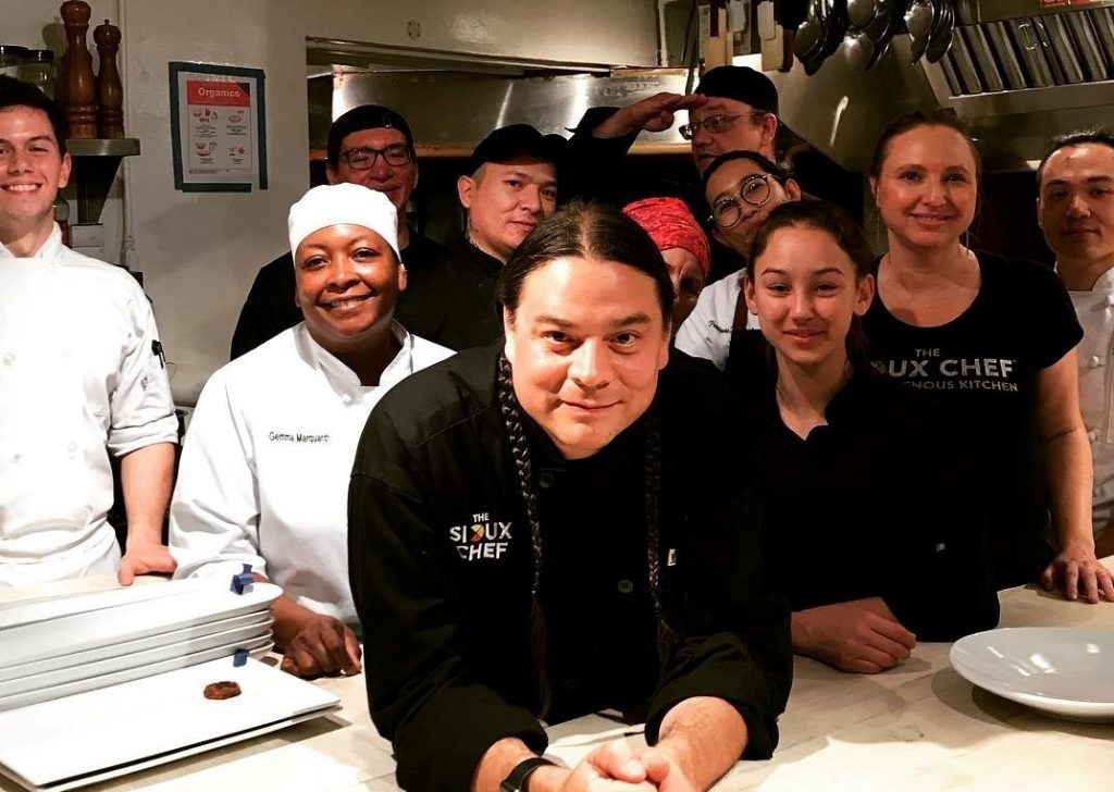 The Sioux Chef leans over a kitchen counter with a team of cooks behind him