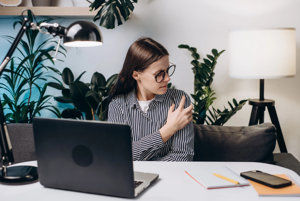 Woman with glasses sits at a desk in a plant-filled home office, adjusting her striped sleeve beside a laptop.