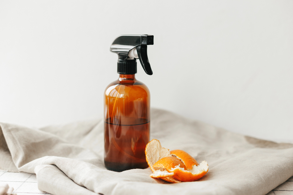 Amber glass spray bottle with a black nozzle sits on a light cloth, with orange peels nearby, suggesting citrus cleaner.